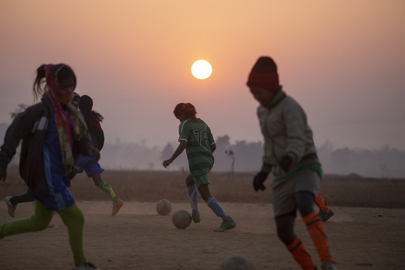 Girls in football kit dribbling balls on a dusty ground at sunrise in Jharkhand, India