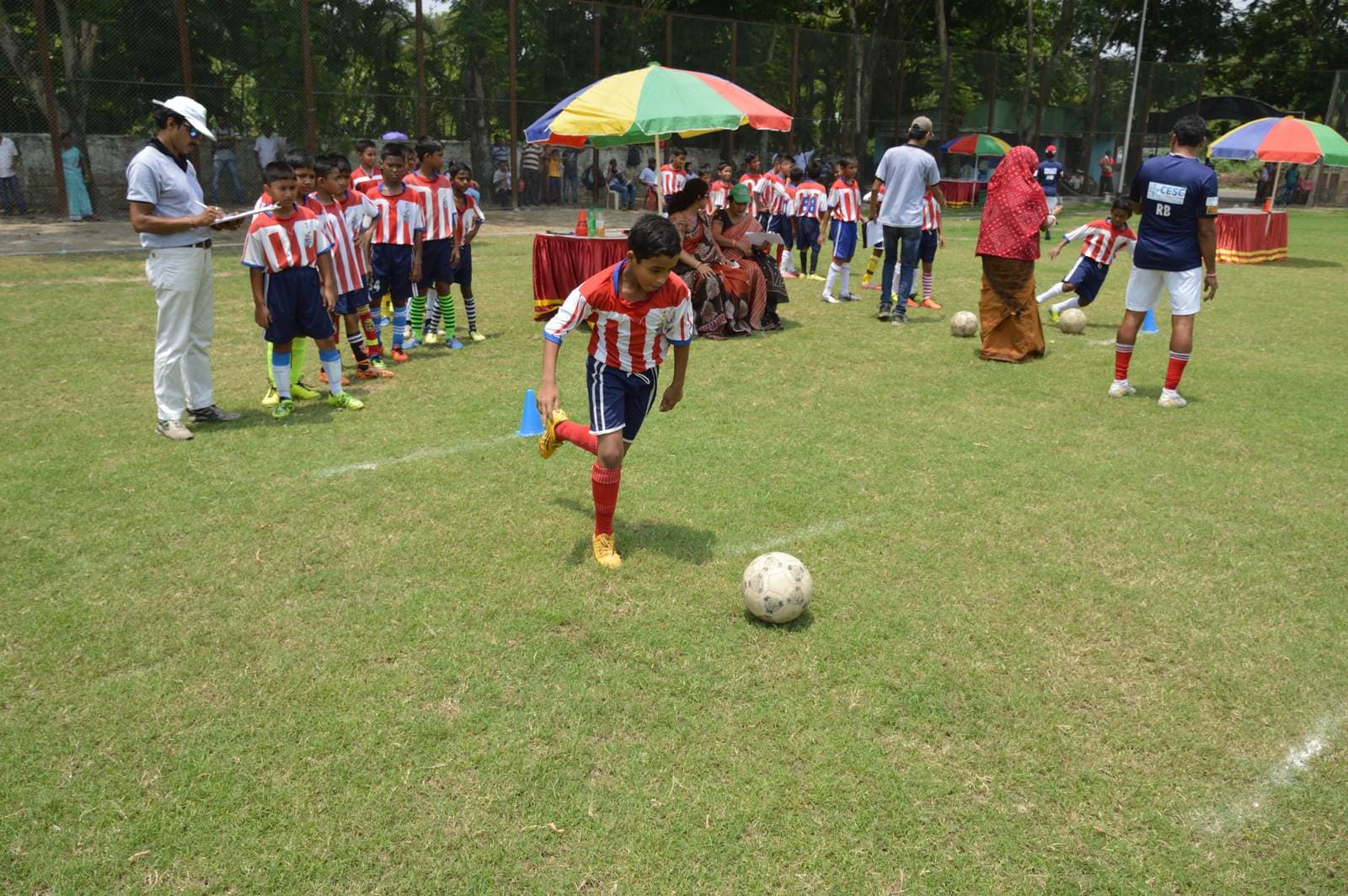 Under-14 boy in a red-and-white kit dribbles a football on grass while teammates watch at a Kolkata grassroots session