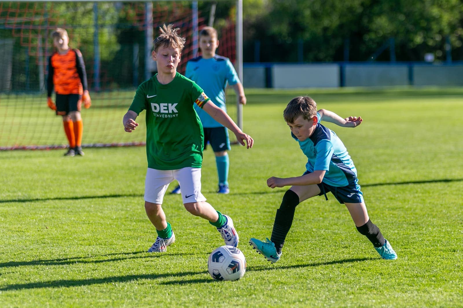 Two young footballers contesting a dribble on a grass pitch with a goalkeeper behind