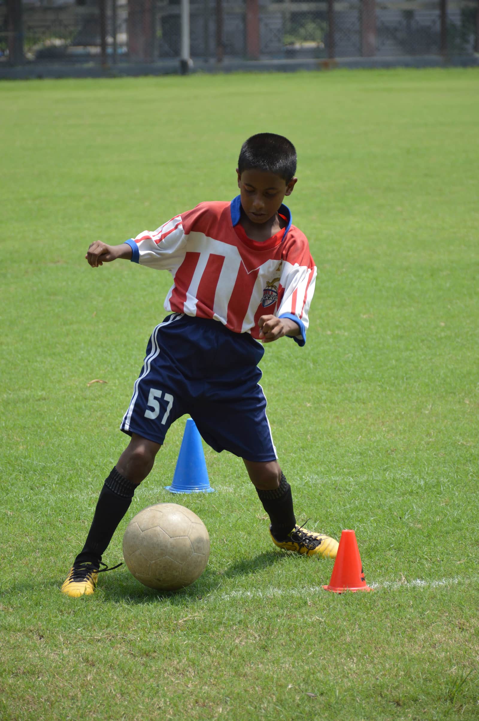 Young footballer in a red-and-white kit controls a ball between cones at an under-14 session in Kolkata