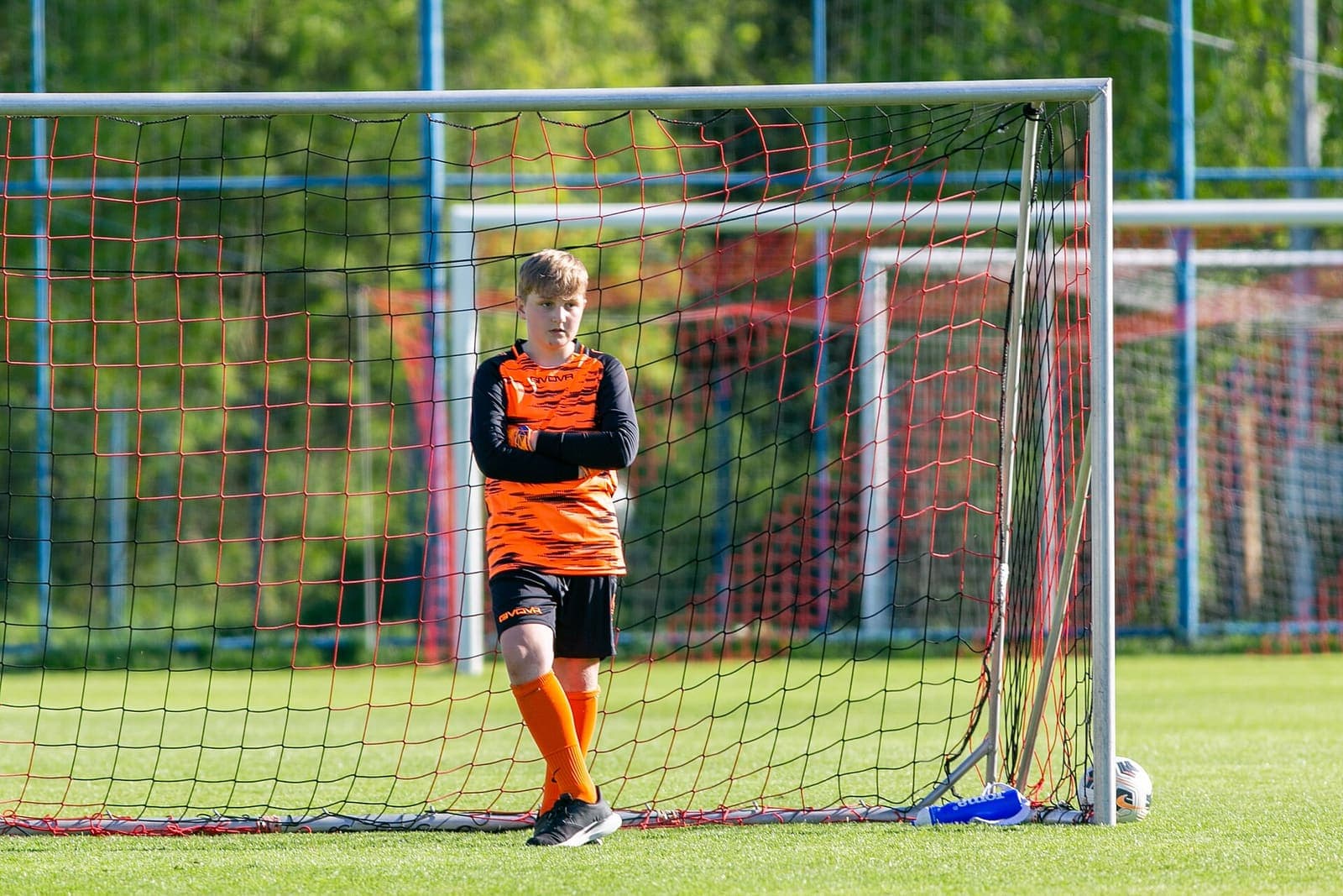Young goalkeeper in orange kit standing in the goal with a football by the post